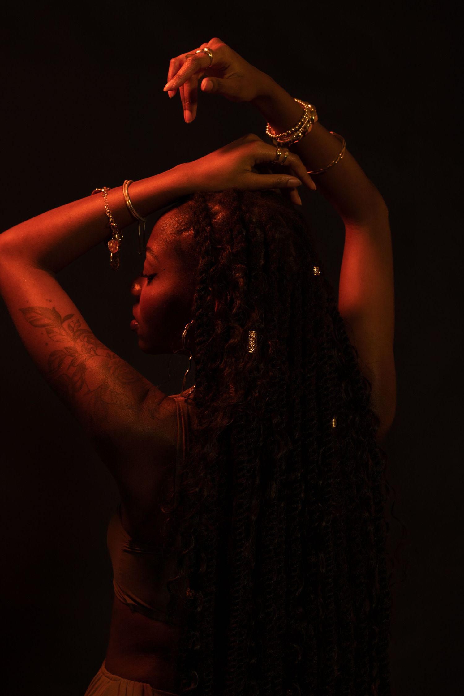 Back view of a Black woman with long braids, gold bracelets, and arm tattoo raising hands in warm light.