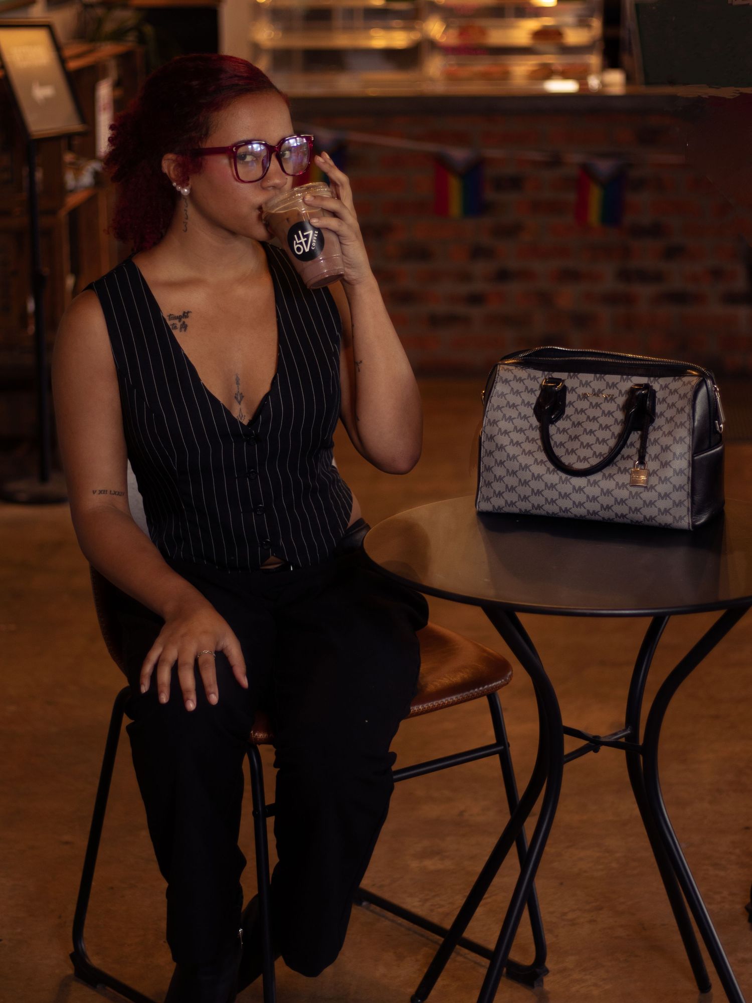 Woman in pinstripe vest sipping a drink at a bar table with a designer handbag.