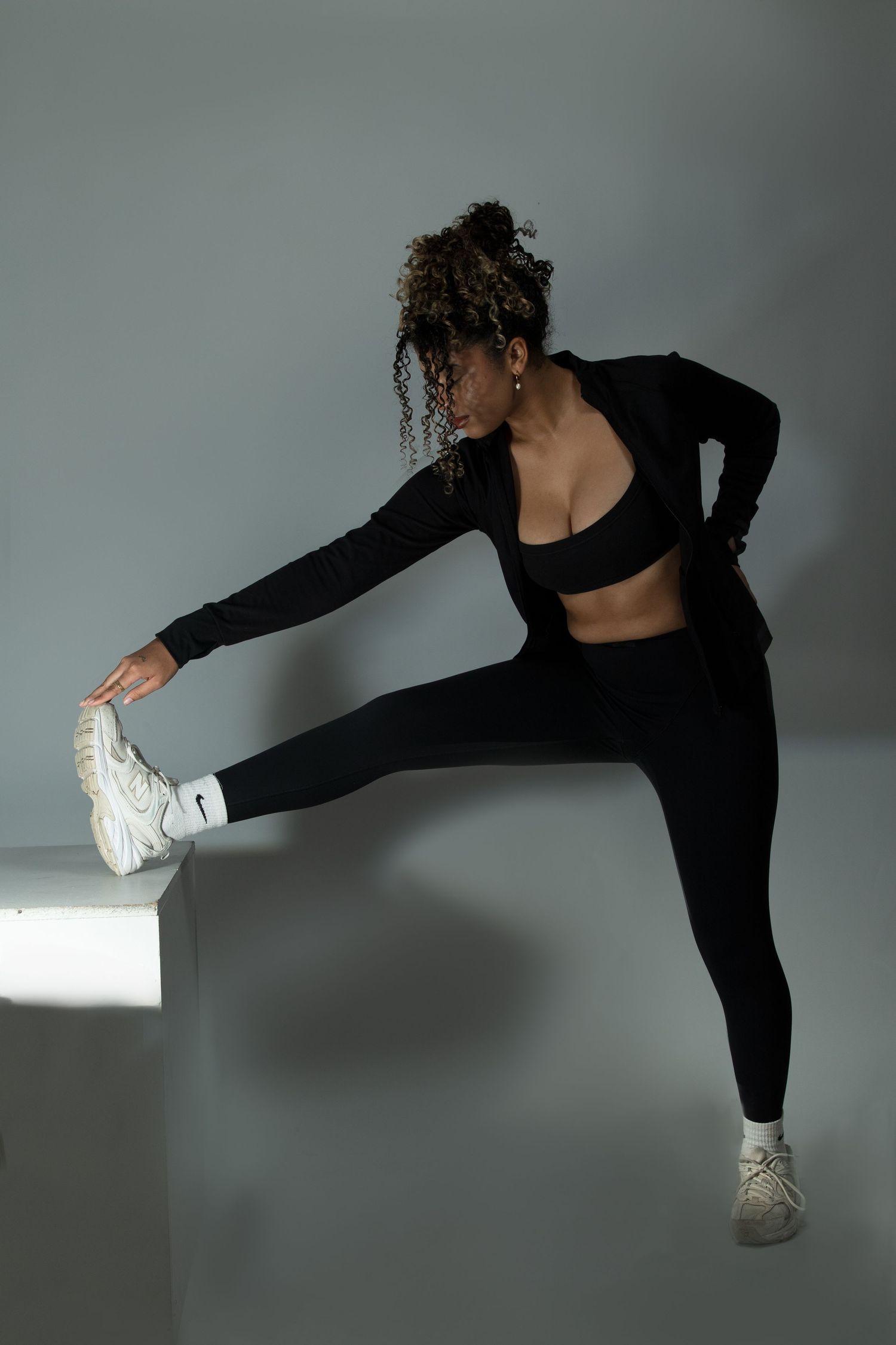 Athletic woman in black sports outfit stretches her leg on a white cube in a studio setting.