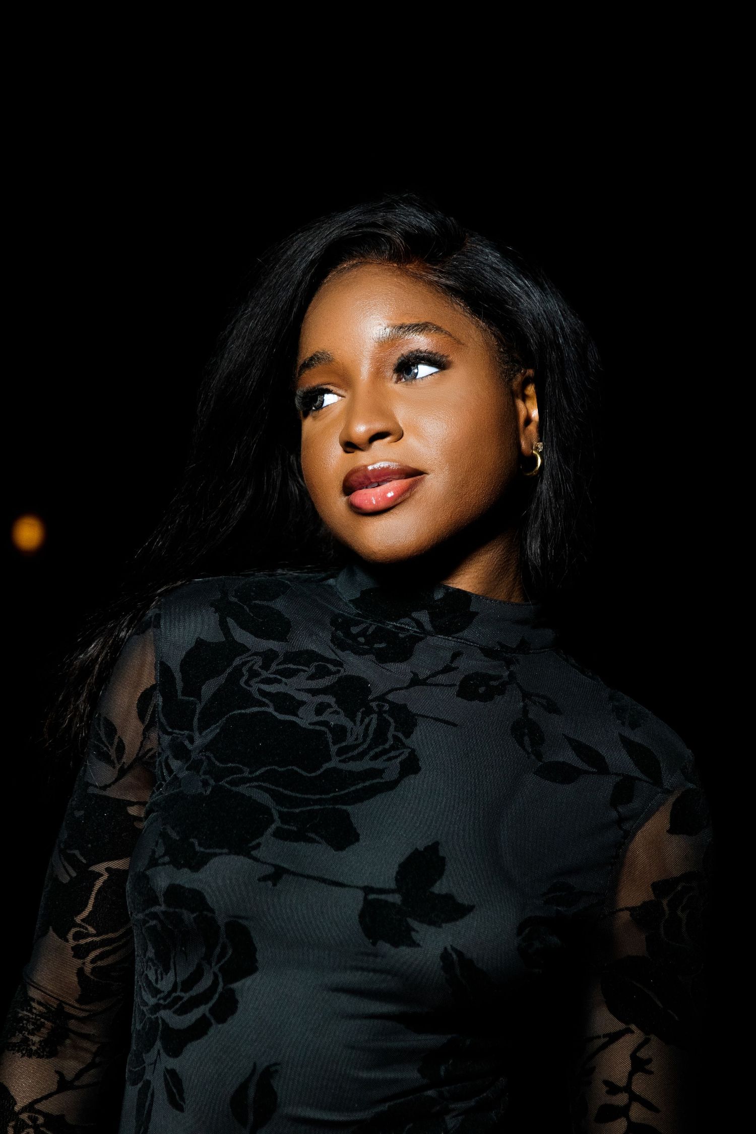 Young Black woman with long straight hair gazing upward in a black floral dress at night.