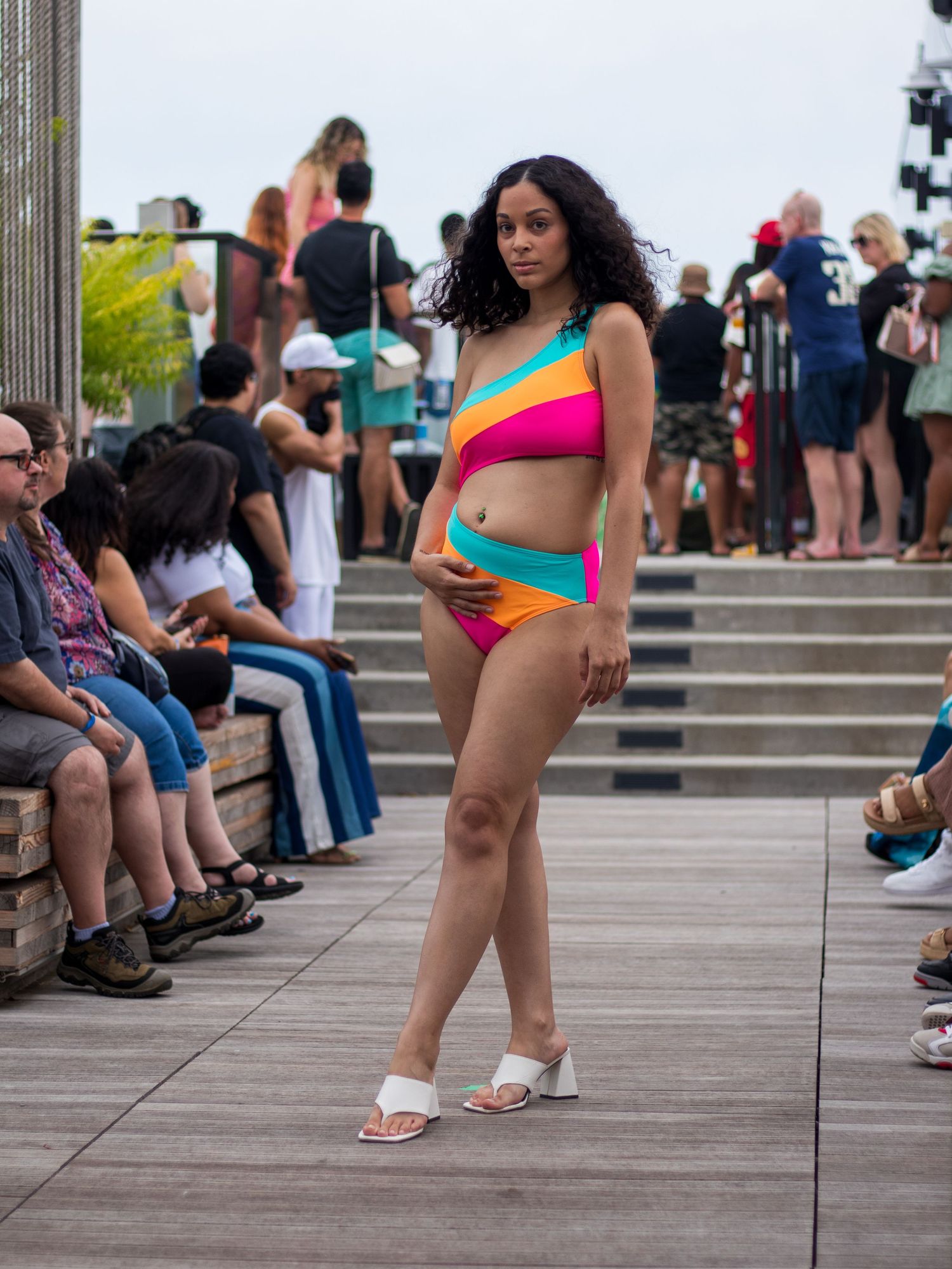 Model in colorful striped bikini set walks outdoor runway show on boardwalk.