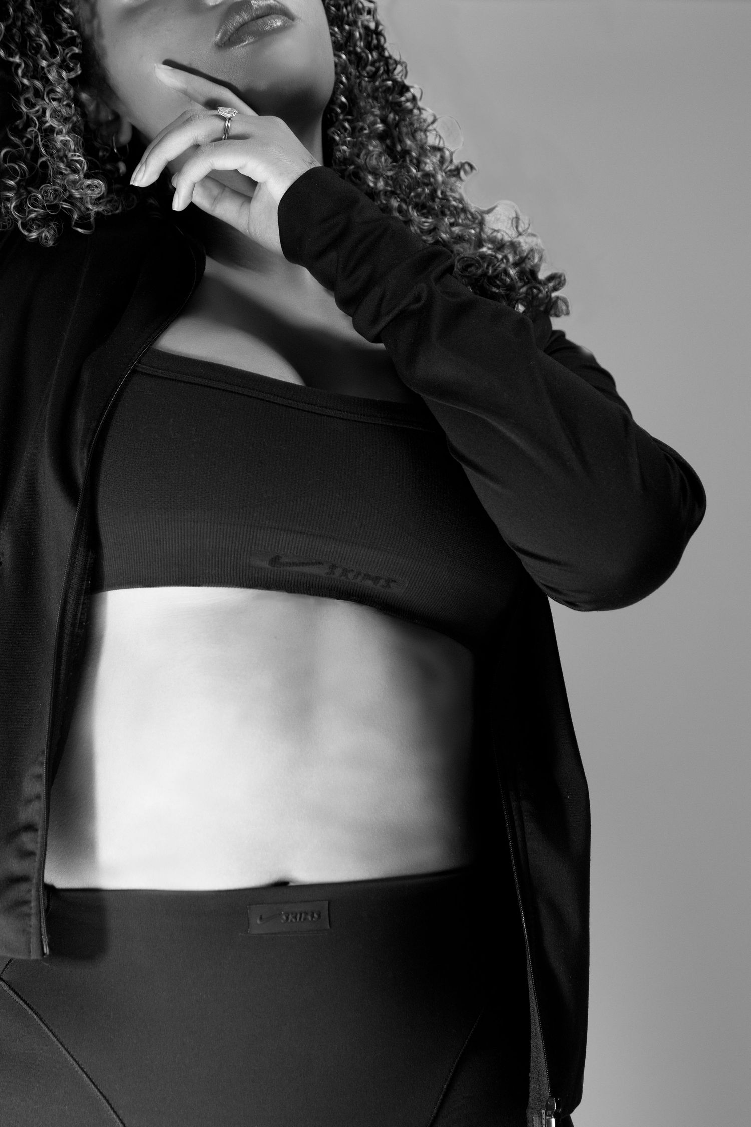 Black and white close-up of a curly-haired woman in a black sports bra and open jacket athleisure outfit.