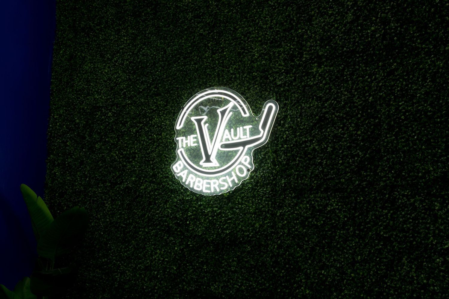 Glowing neon sign for The Vault Barbershop mounted on a dark green artificial grass wall.