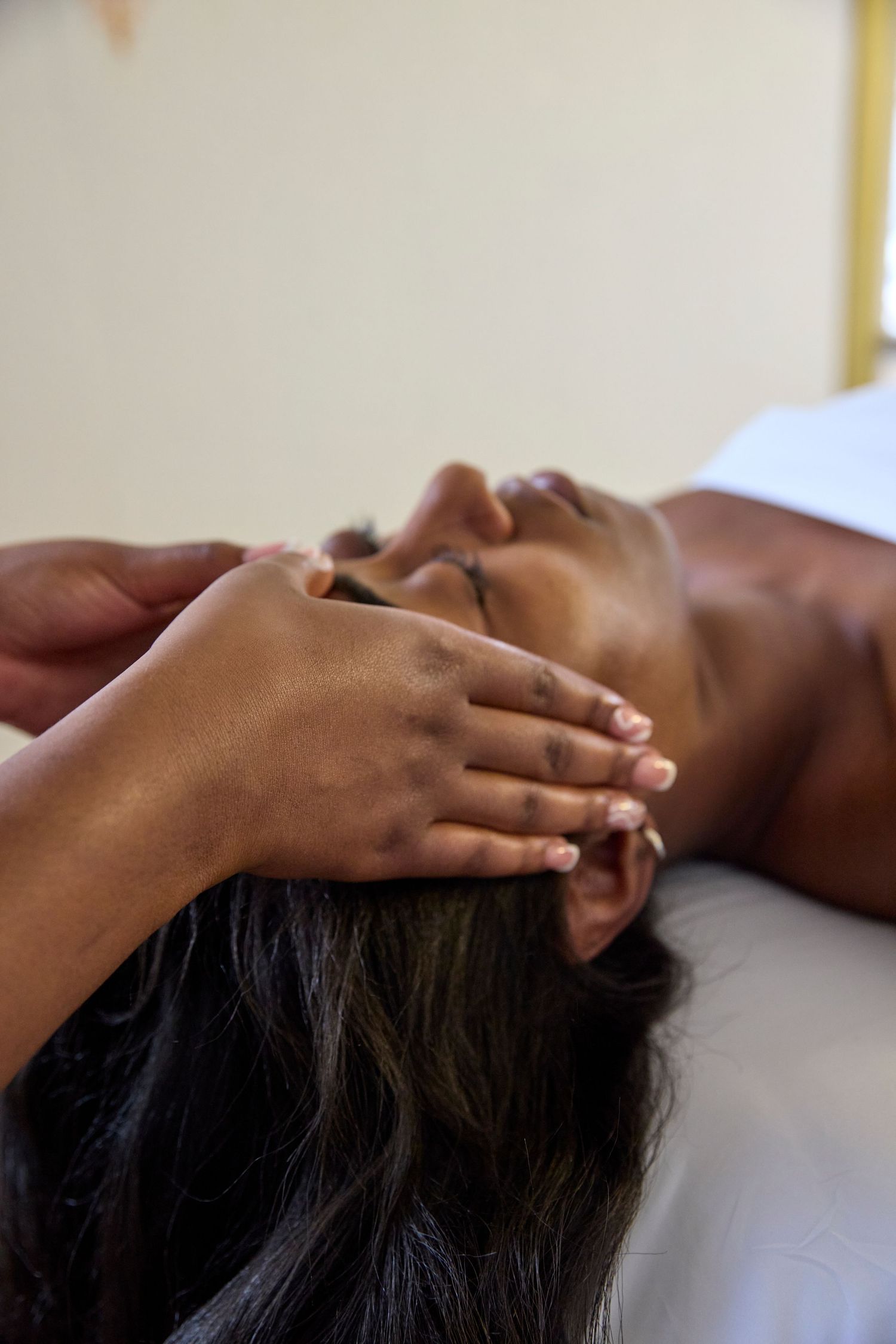 A woman receiving a relaxing head massage at a spa, lying on a white massage table with eyes closed.