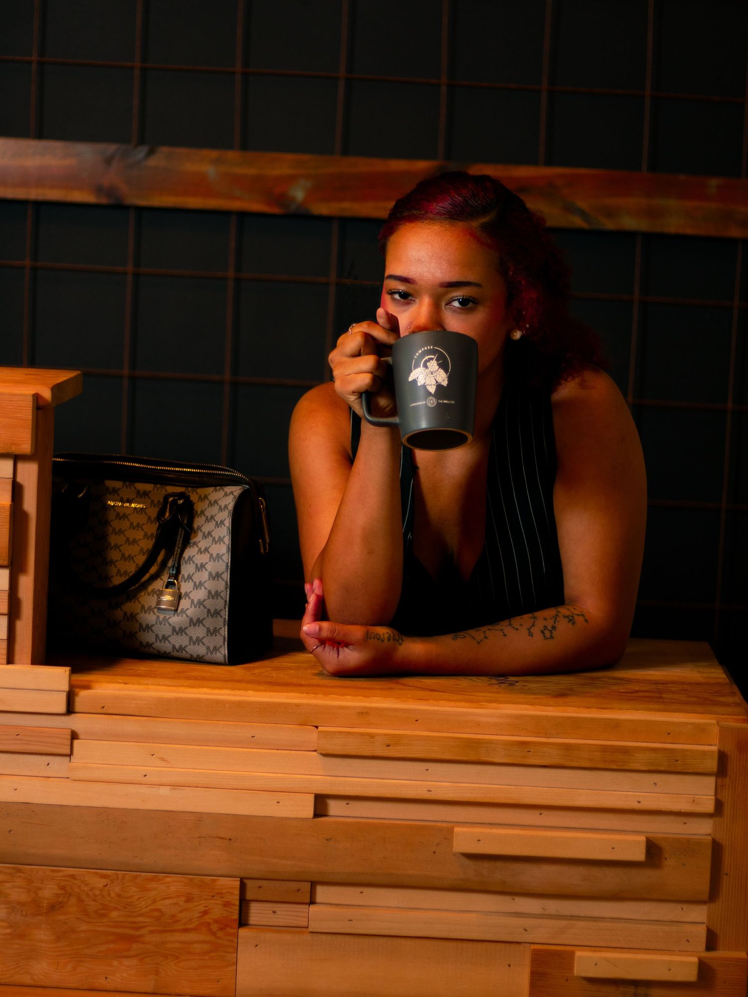 Woman drinking from dark ceramic mug at wooden table with designer bag in cafe.
