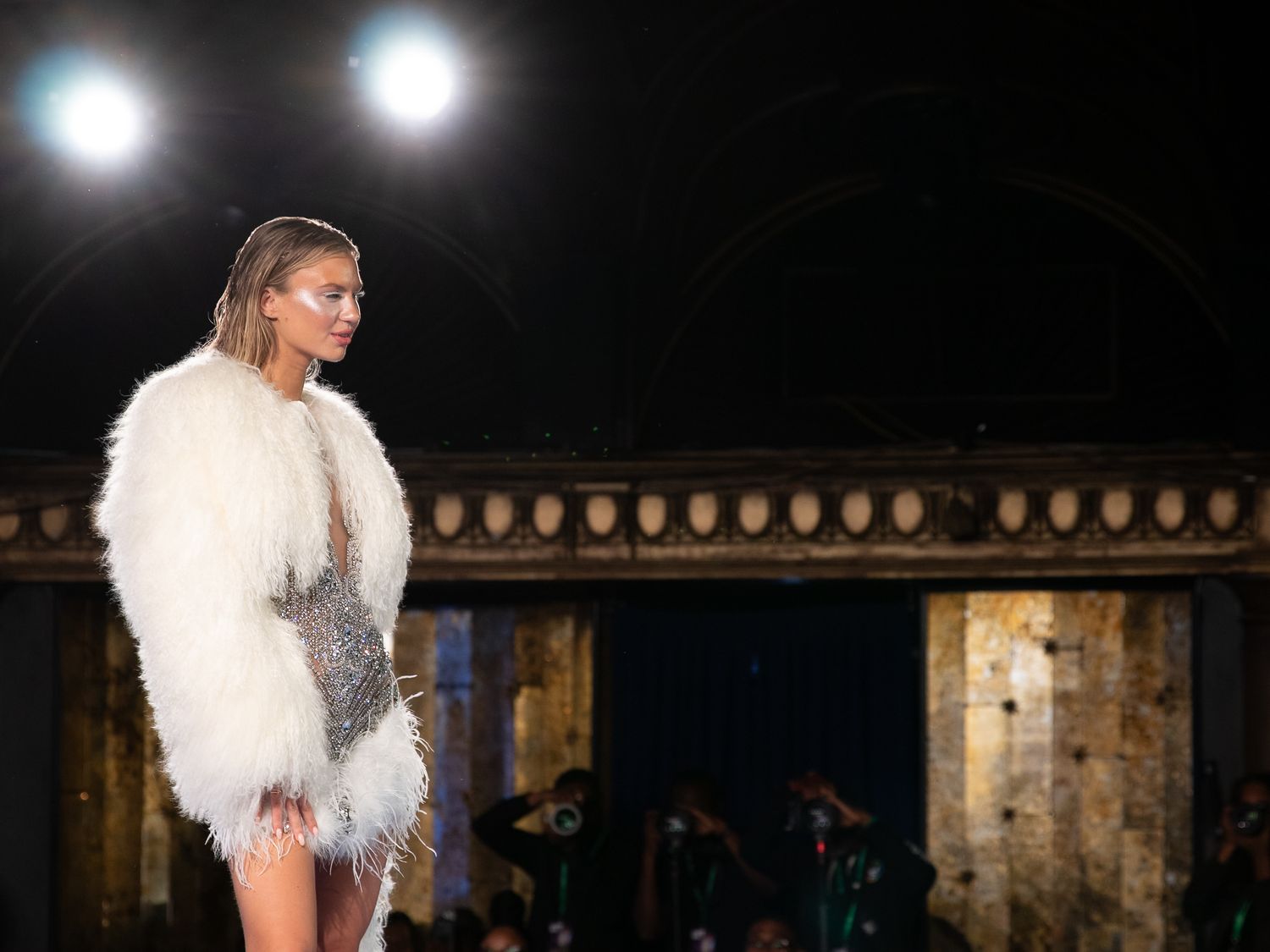 Model walks runway in white fluffy fur coat with embellished mini dress at fashion show.
