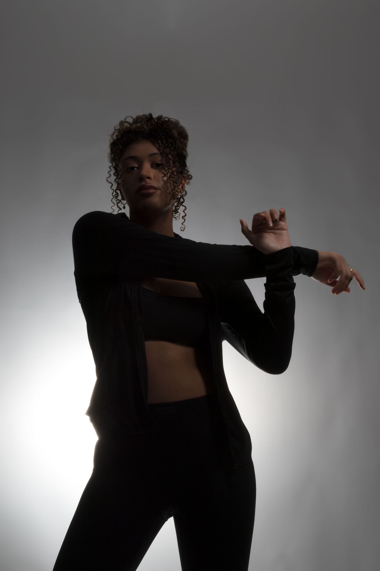 A woman in black athletic wear poses gracefully with raised arms against a bright studio background.