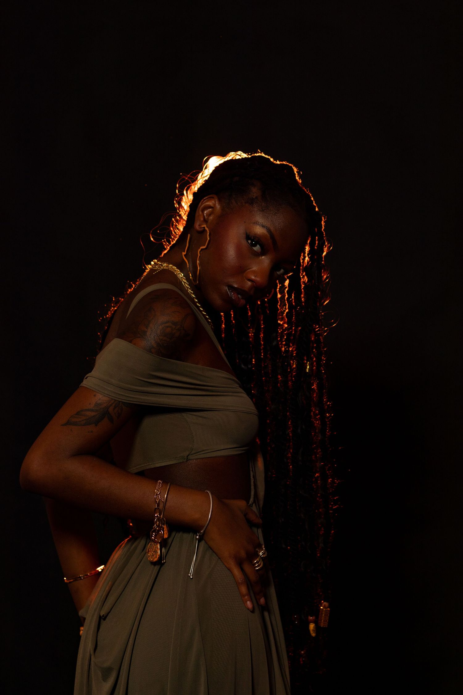 Black woman with long braids, tattoo, gold jewelry, and olive crop top posing against dark background.