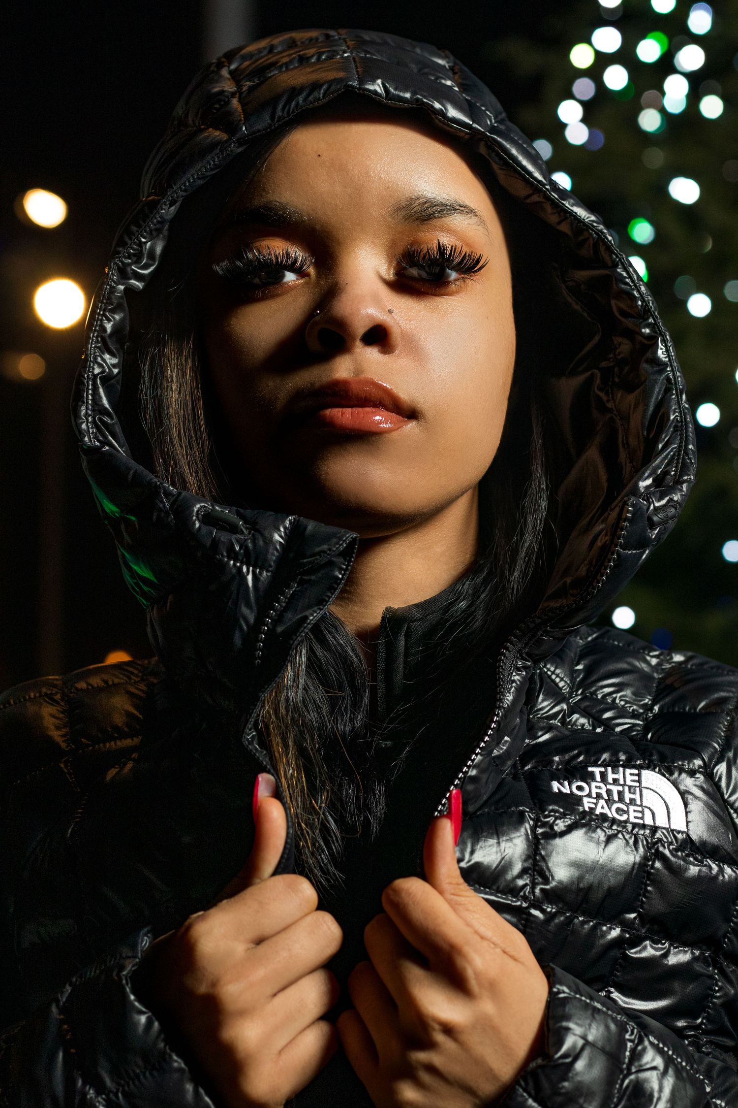 Woman in black North Face puffer jacket with dramatic lashes near holiday Christmas tree lights.