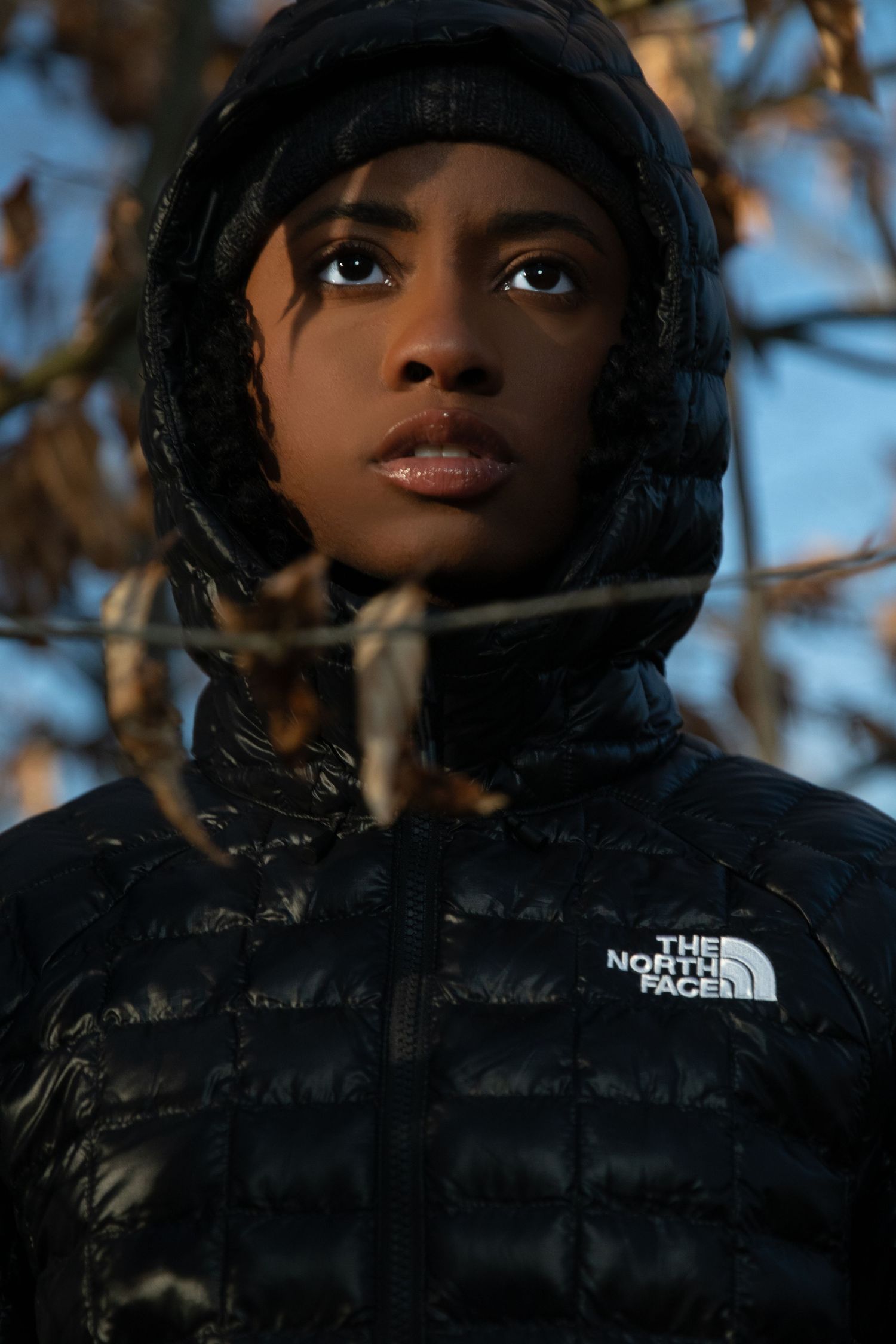 Woman in black The North Face hooded puffer jacket outdoors among autumn branches.