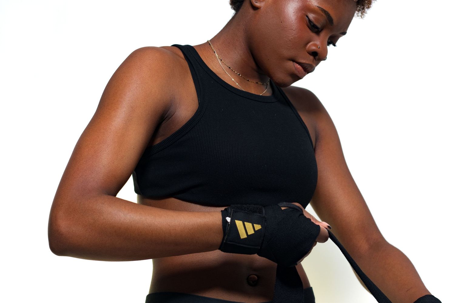 Athletic Black woman in black sports bra wrapping hands with Adidas boxing wraps on white background.