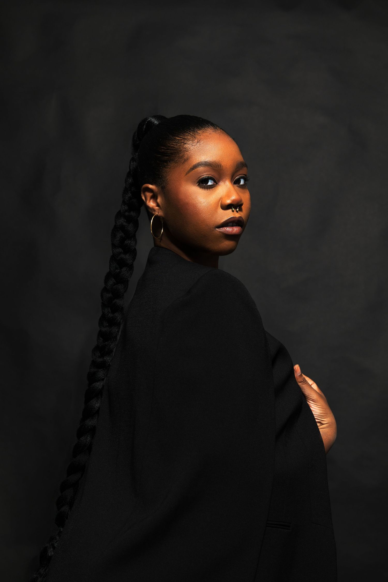 Woman in black blazer with long braided ponytail posing against dark studio background.