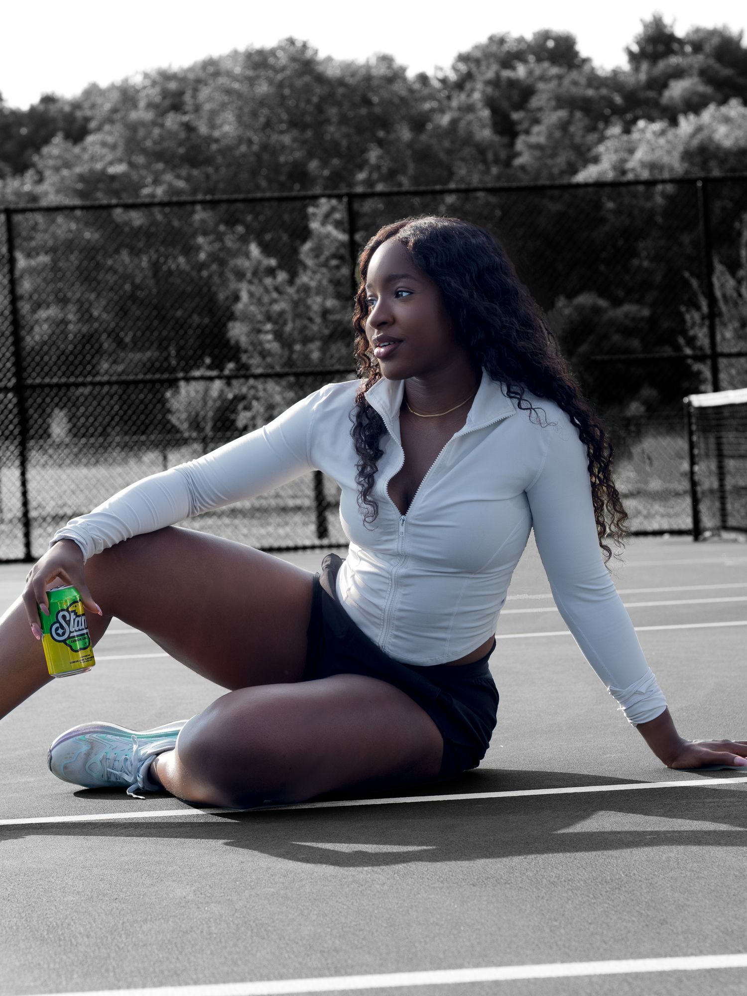 Young woman in white zip-up top and black shorts sitting on a tennis court holding a Starry soda can.