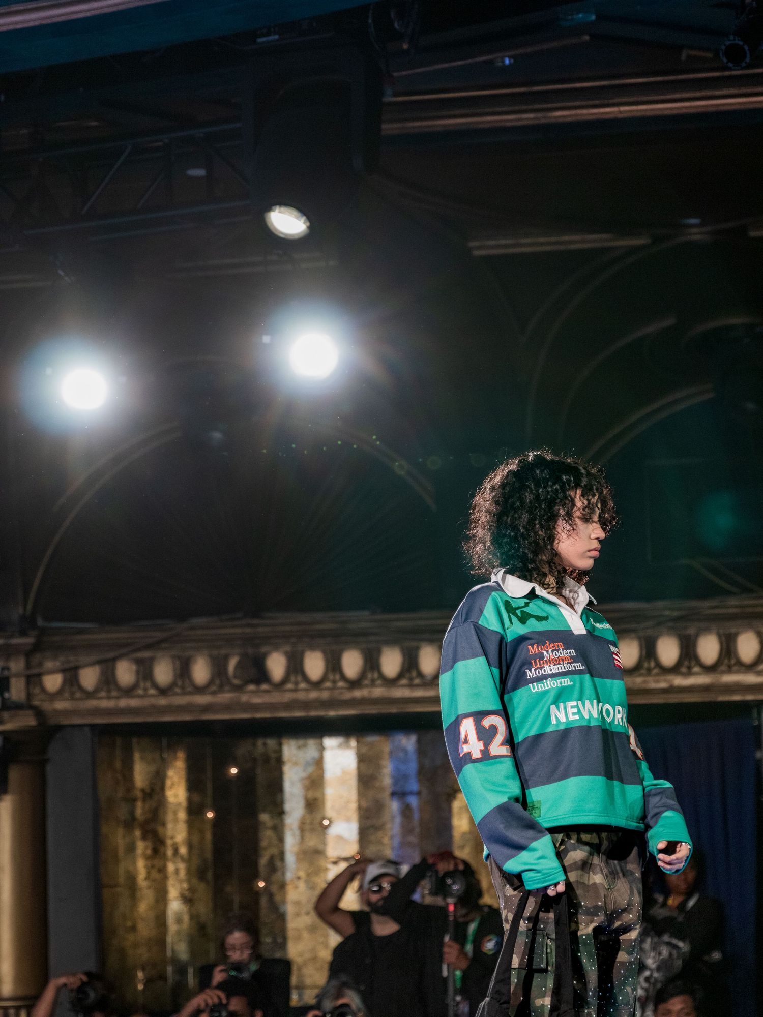 Model wearing oversized green striped rugby jersey numbered 42 on a dramatic lit fashion runway stage.
