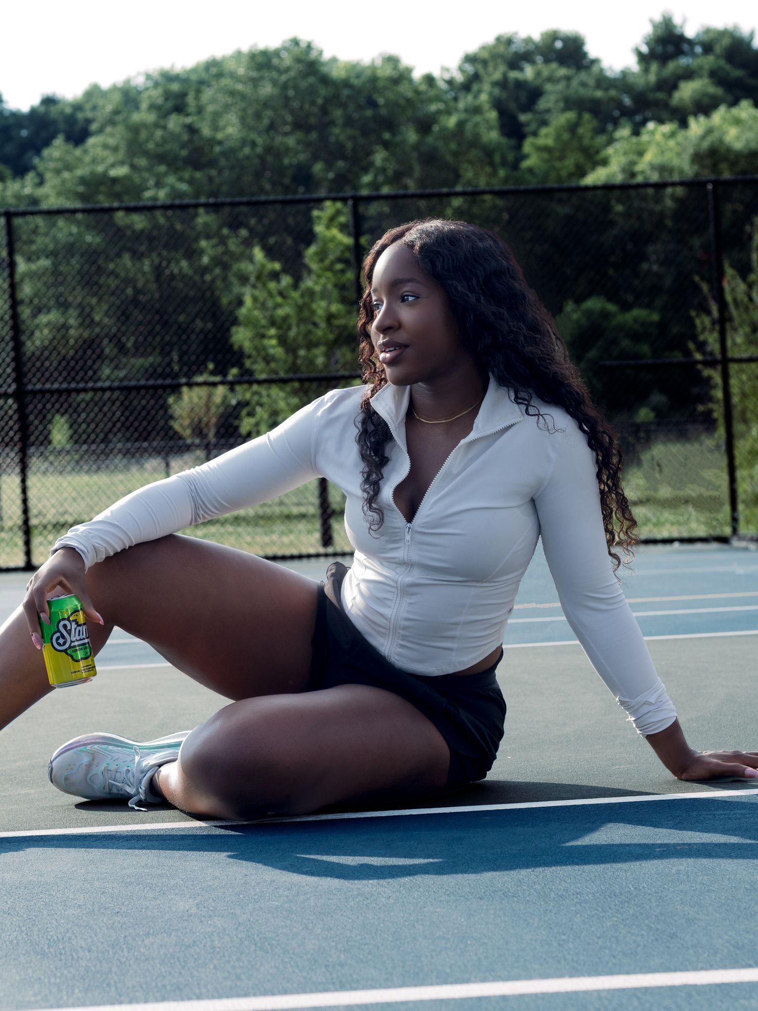 Young woman in white athletic top sitting relaxed on a tennis court holding a green soda can outdoors.