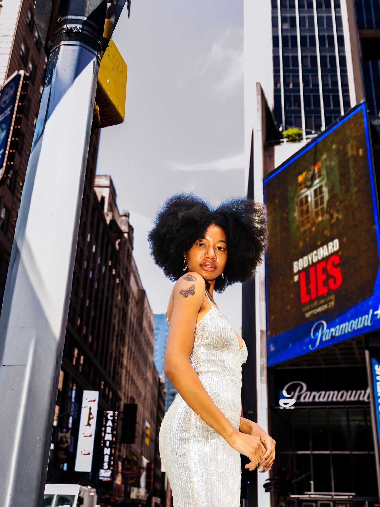 Black woman in silver sequin dress poses on Times Square sidewalk with Paramount billboard in background.