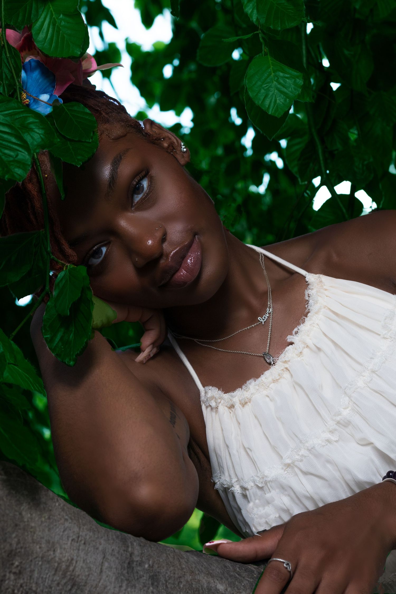 Woman in white dress rests against tree trunk surrounded by green leaves in dappled sunlight.
