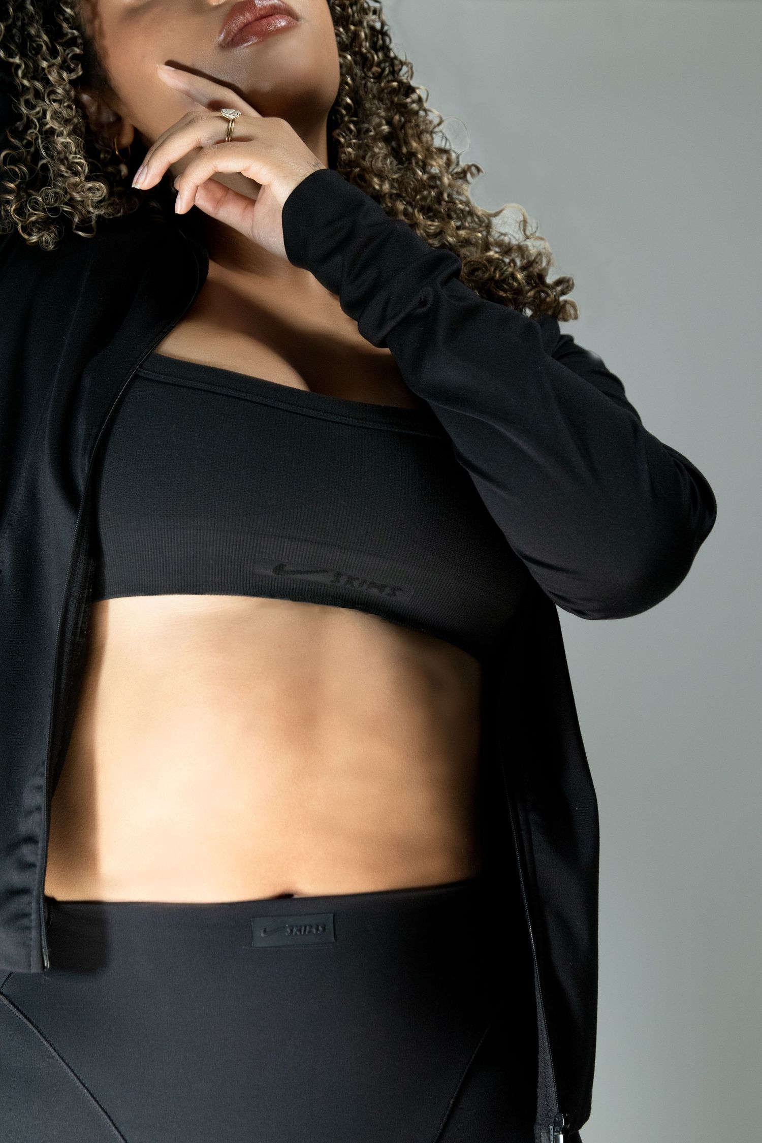 Close-up of a curly-haired woman in black sports bra and athletic jacket showing midriff in studio.
