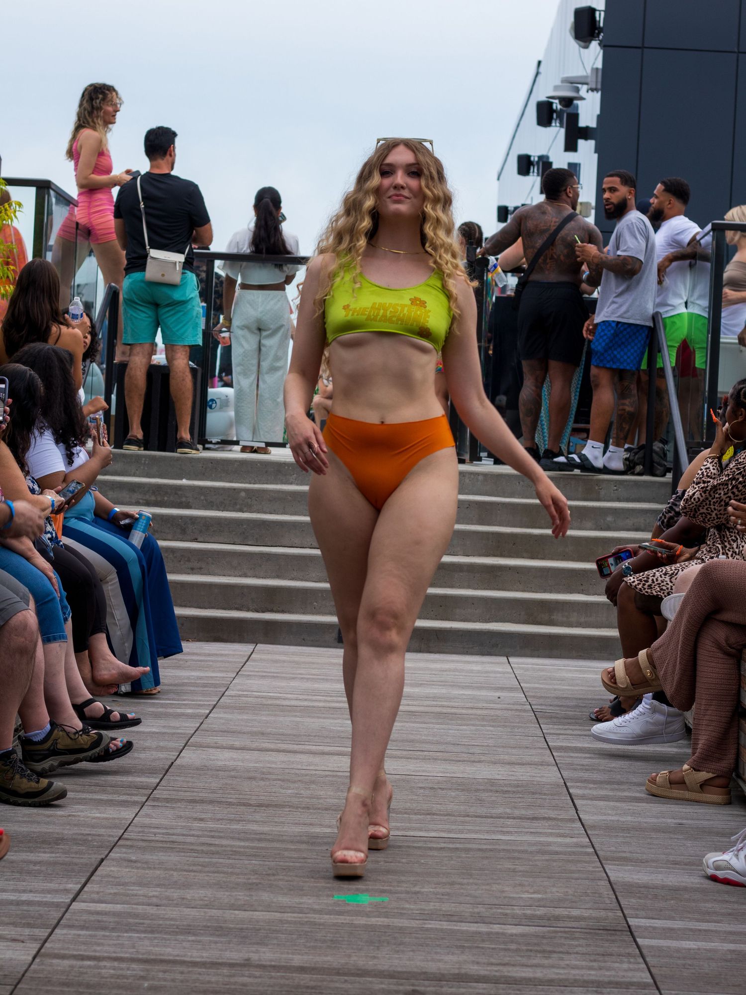 Curly-haired model walks runway in green crop top and orange bikini bottoms with platform heels.