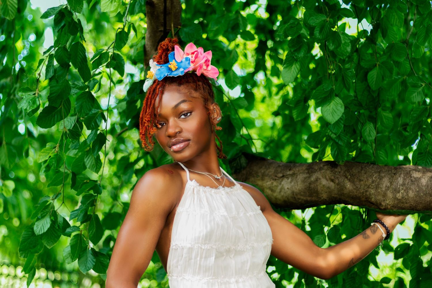 Beautiful woman wearing floral headpiece leans against tree trunk surrounded by vibrant green foliage.