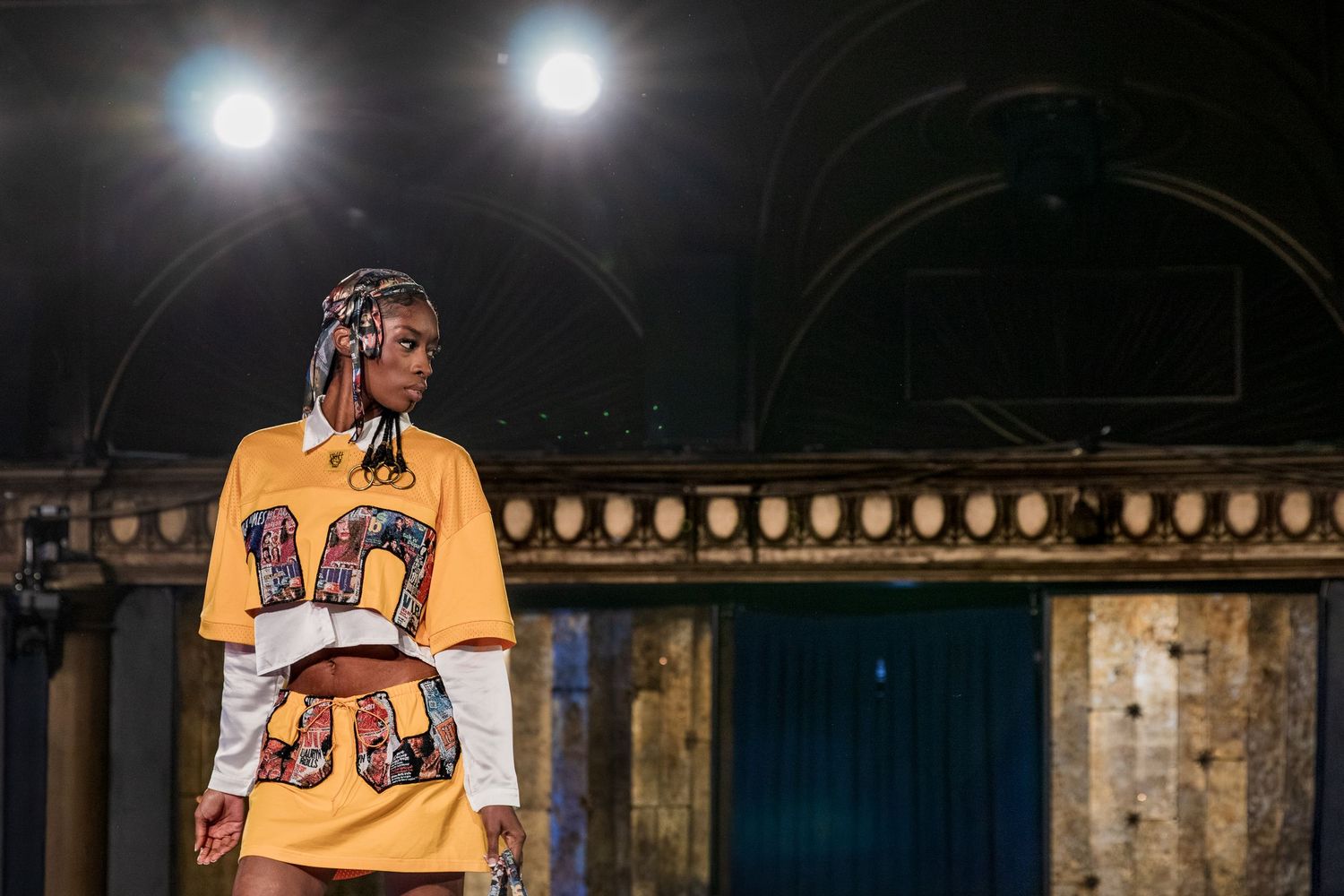 Model wears yellow varsity-style graphic dress over white long sleeve shirt on a fashion show runway.