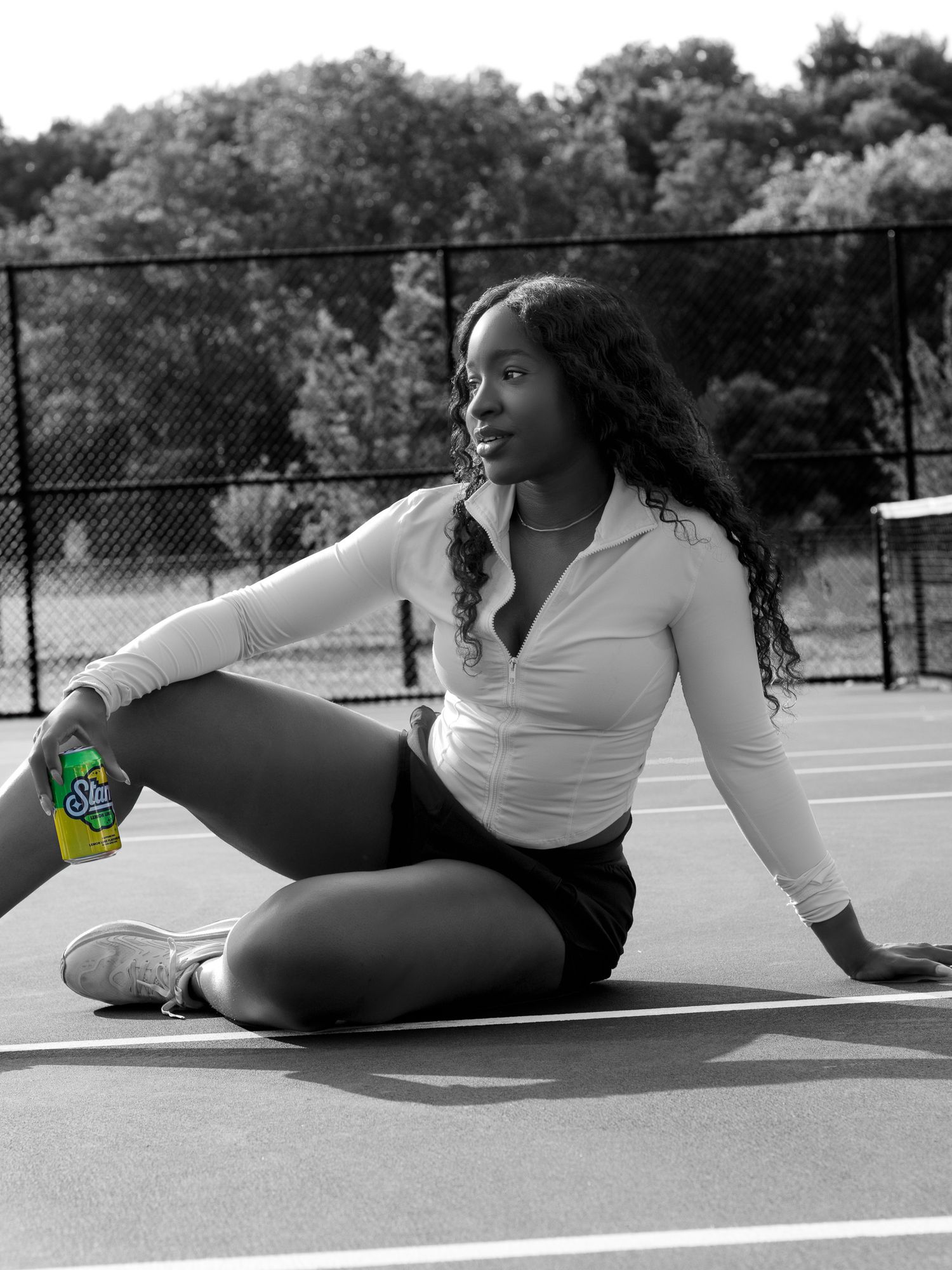 Black and white photo of a young woman sitting on a tennis court holding a bright yellow-green soda can.