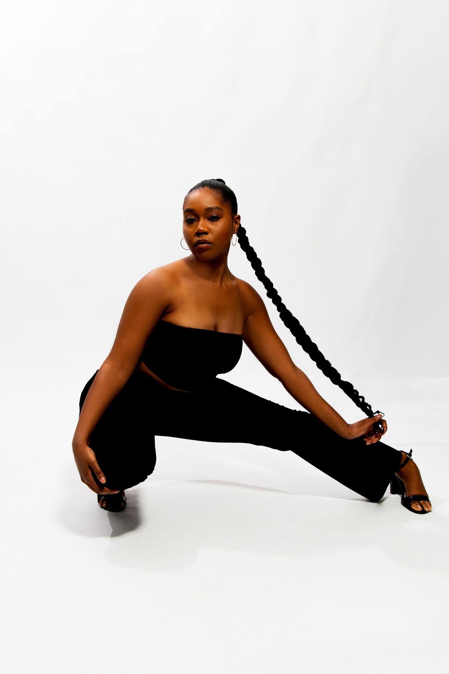 Young Black woman in black outfit crouching on floor holding her long braid in a studio setting.