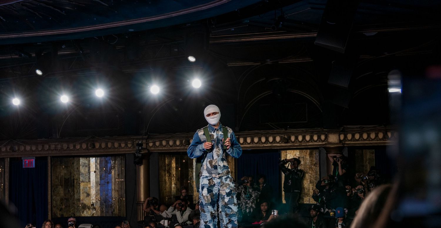 Performer in floral jumpsuit and white face covering speaks on dimly lit stage.