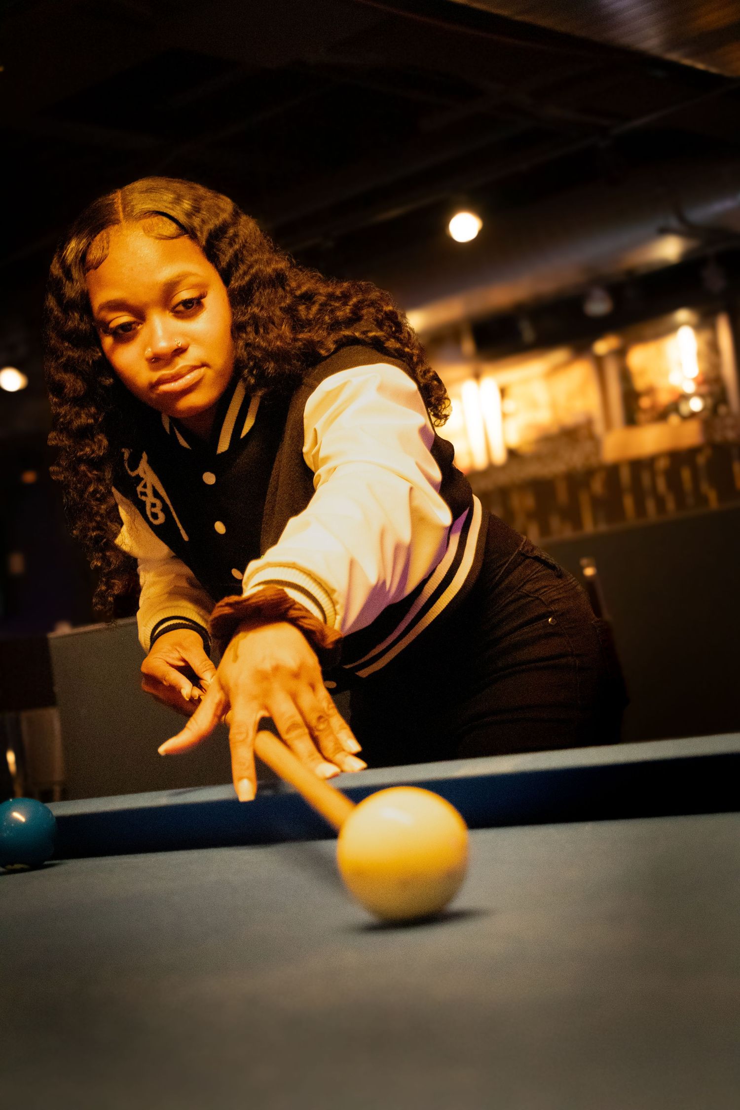 Young woman in varsity jacket shooting pool at a billiard table in a dark bar.