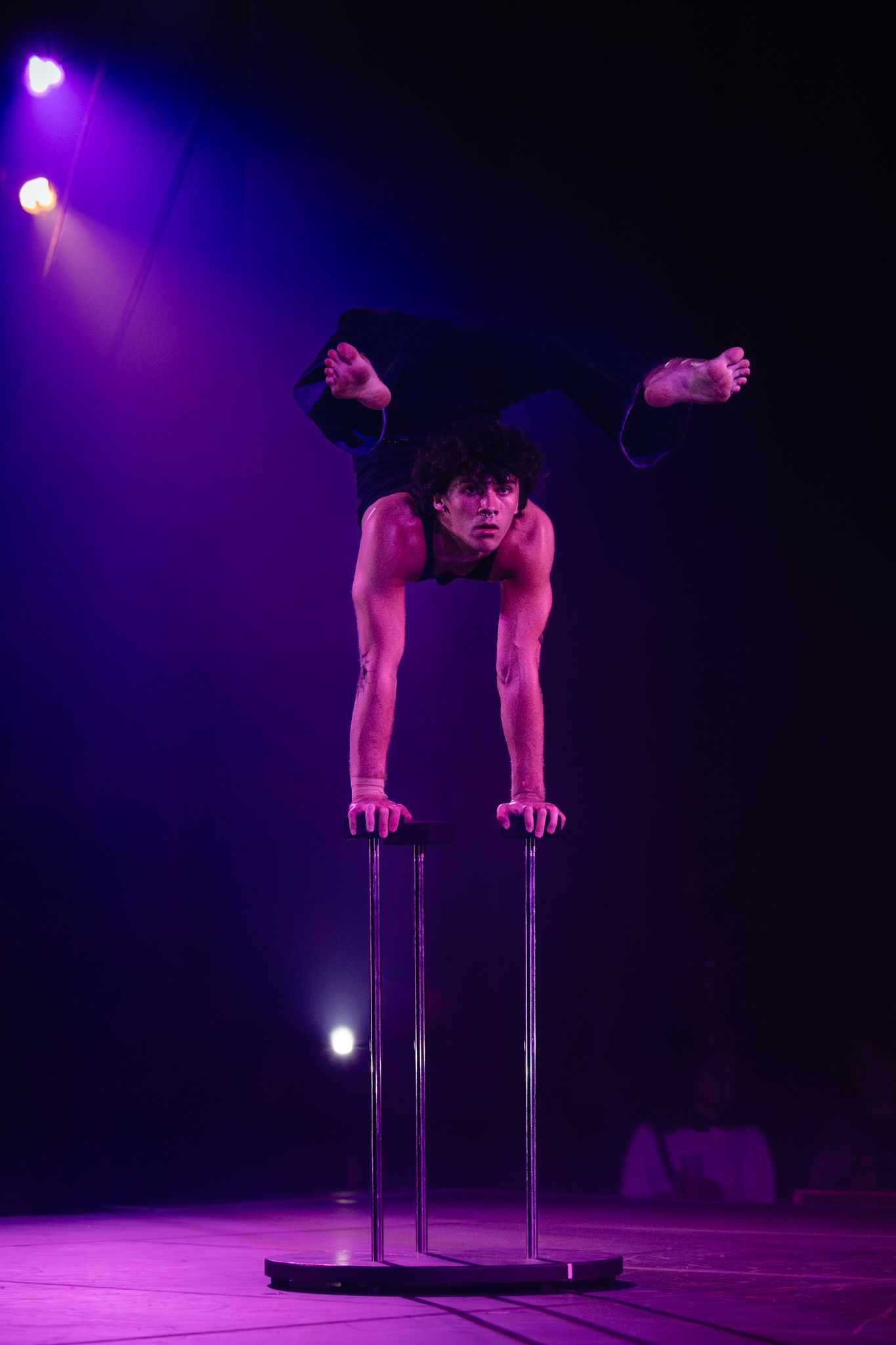Performer in dramatic stage lighting executes strength-based acrobatic pose on parallel bars during circus act.
