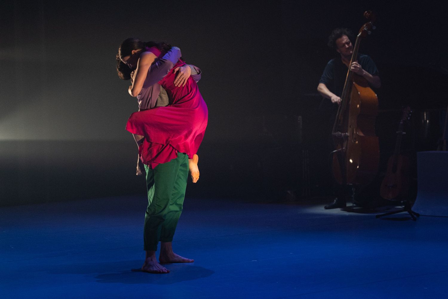 Dancers perform an intimate lift on a blue-lit stage while a bassist plays in the background during a contemporary performance.