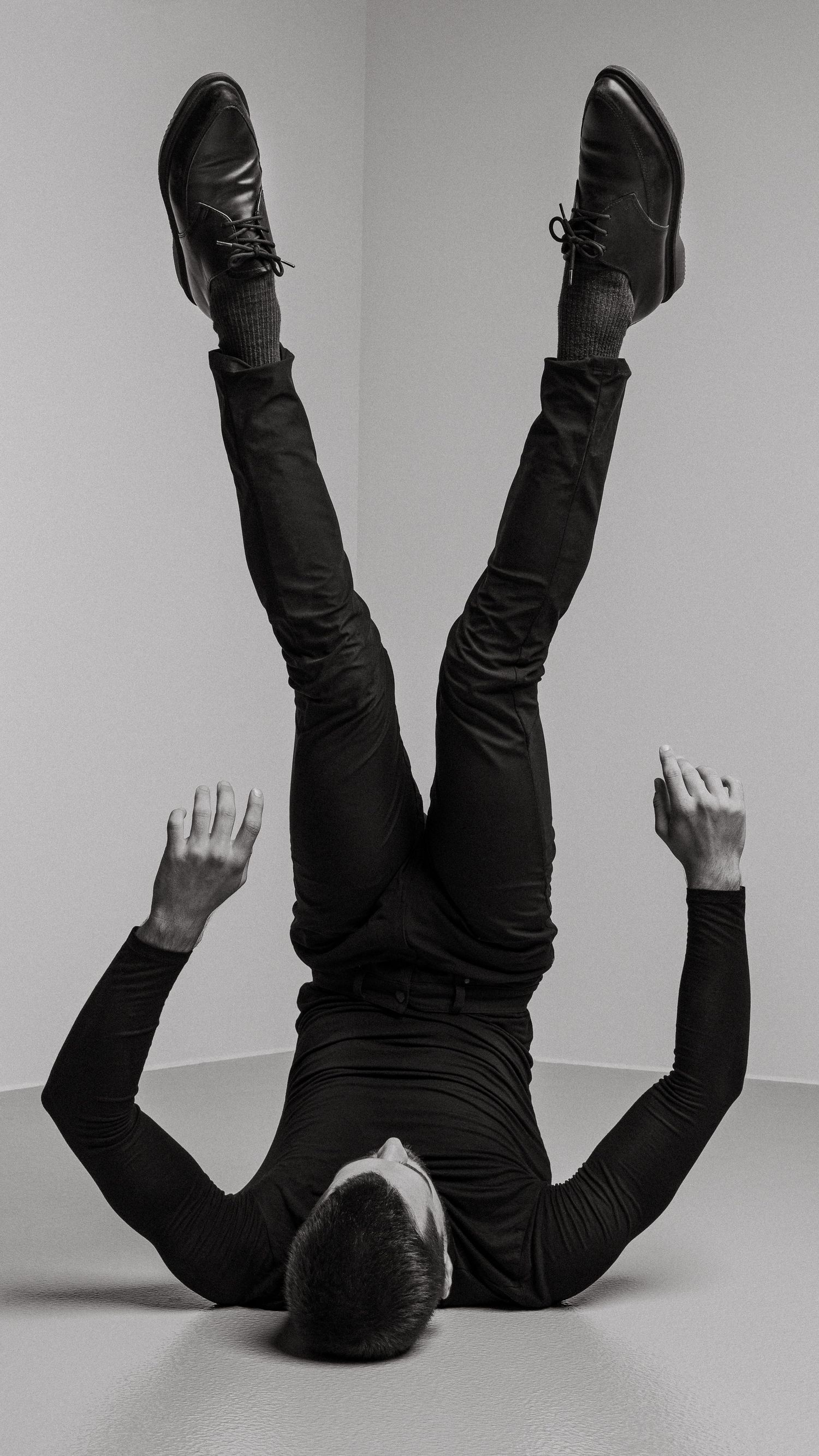 Black and white artistic portrait of person lying upside down against wall wearing formal attire and dress shoes.