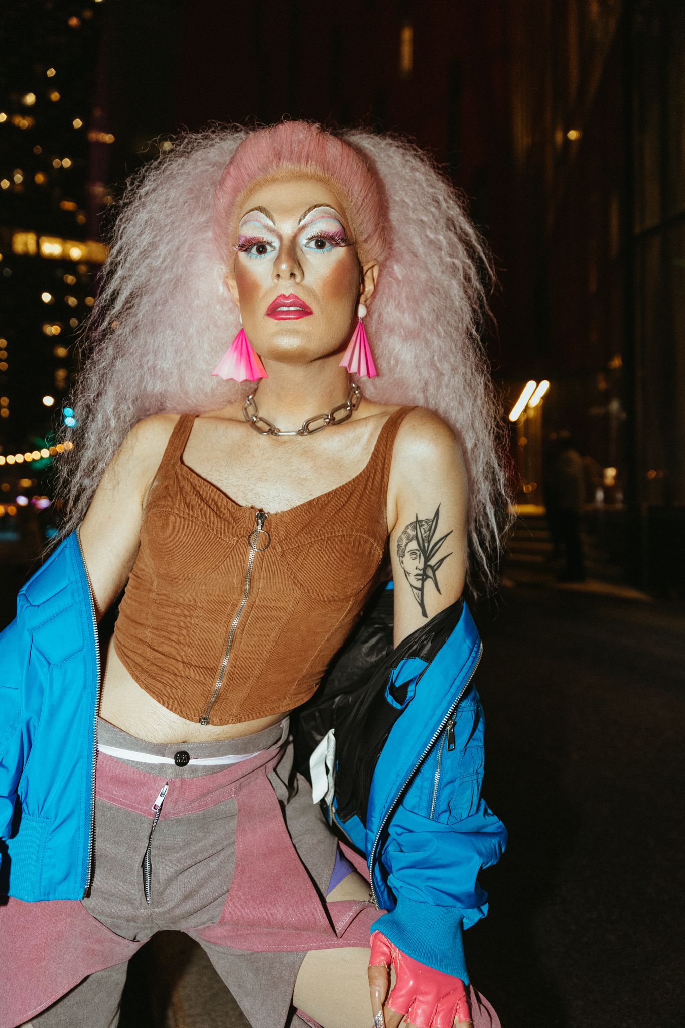 Person with pink hair and bold makeup wearing a brown corset top and blue jacket against city lights at night.