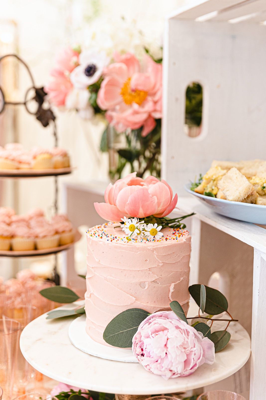 Pink buttercream cake decorated with peonies and sprinkles sits on white cake stand among pastel dessert display.