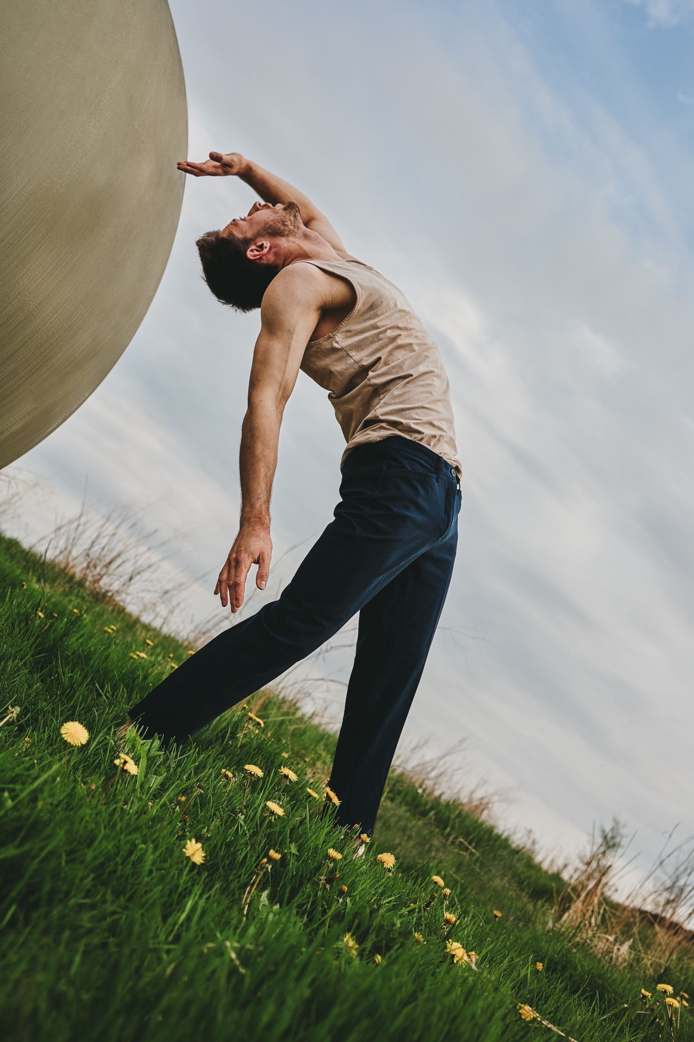 Figure bending and moving with exercise ball on grassy meadow with dandelions under cloudy sky.