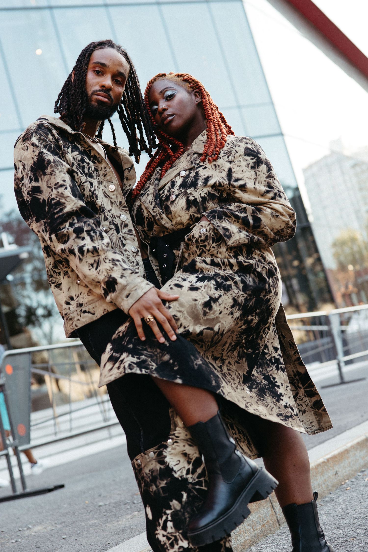 Two people pose together wearing matching brown and beige patterned coats with boots in an urban setting.