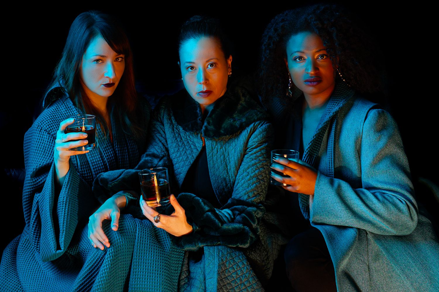 Three figures sit together holding drinks while illuminated in dramatic blue and red lighting.