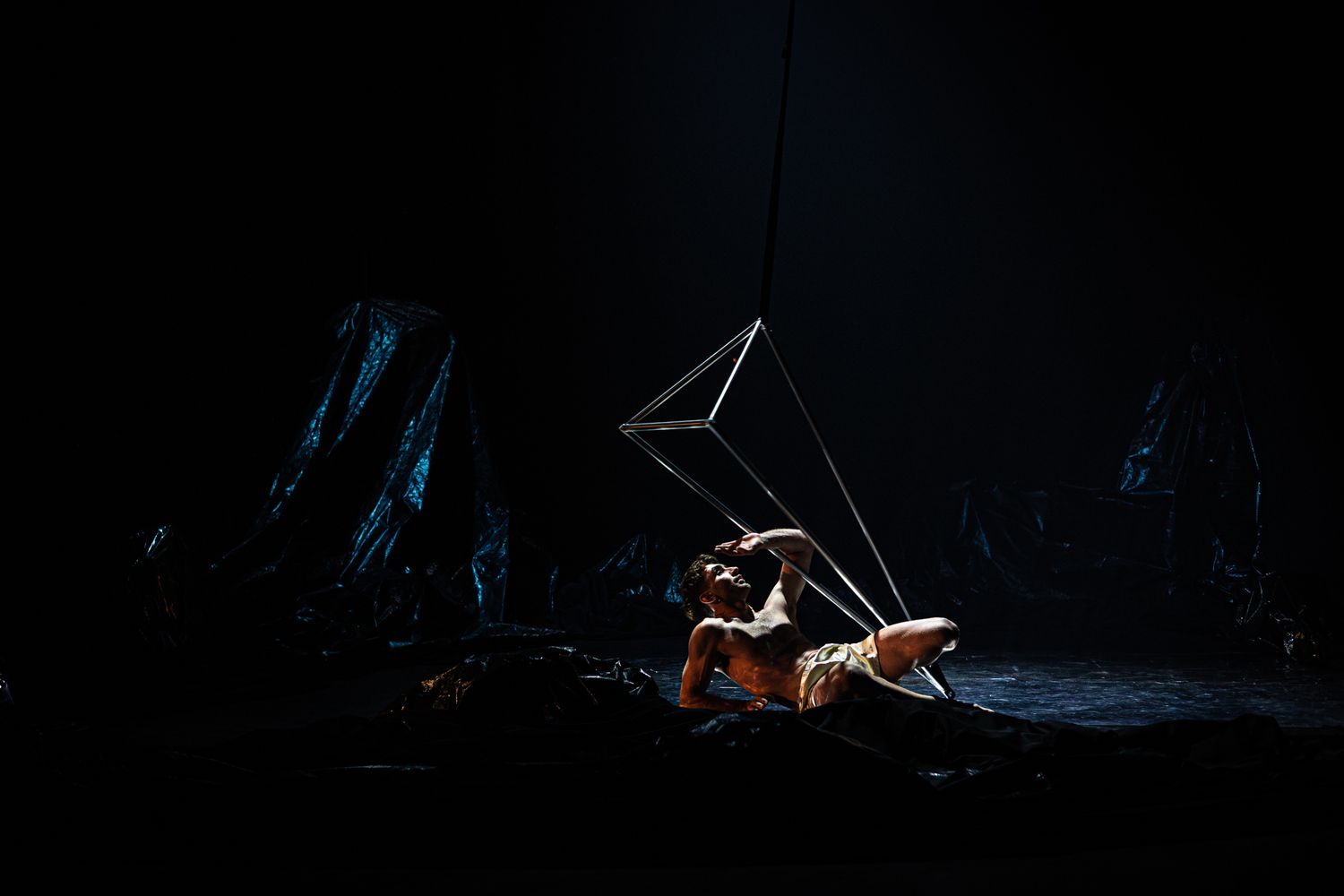 A performer in a sparkly costume executes an aerial acrobatic performance against a dark theatrical backdrop.