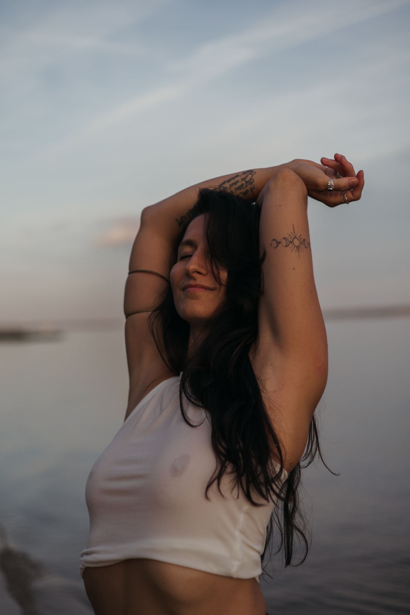 Dark-haired figure in white top poses expressively against a moody waterfront sunset backdrop.