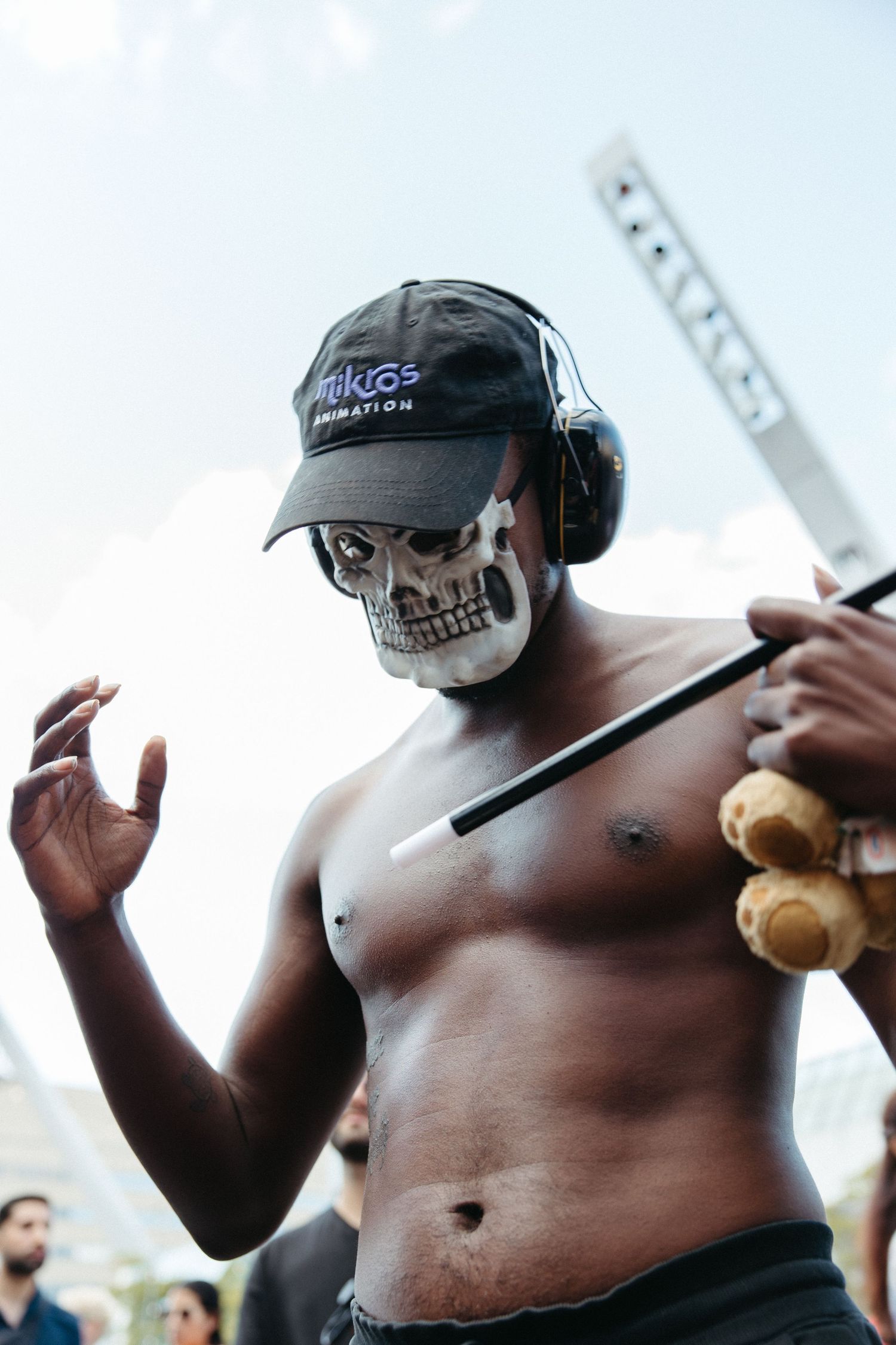 Shirtless festival-goer wearing a skull mask, black cap and headphones gestures while holding a stick at an outdoor event.