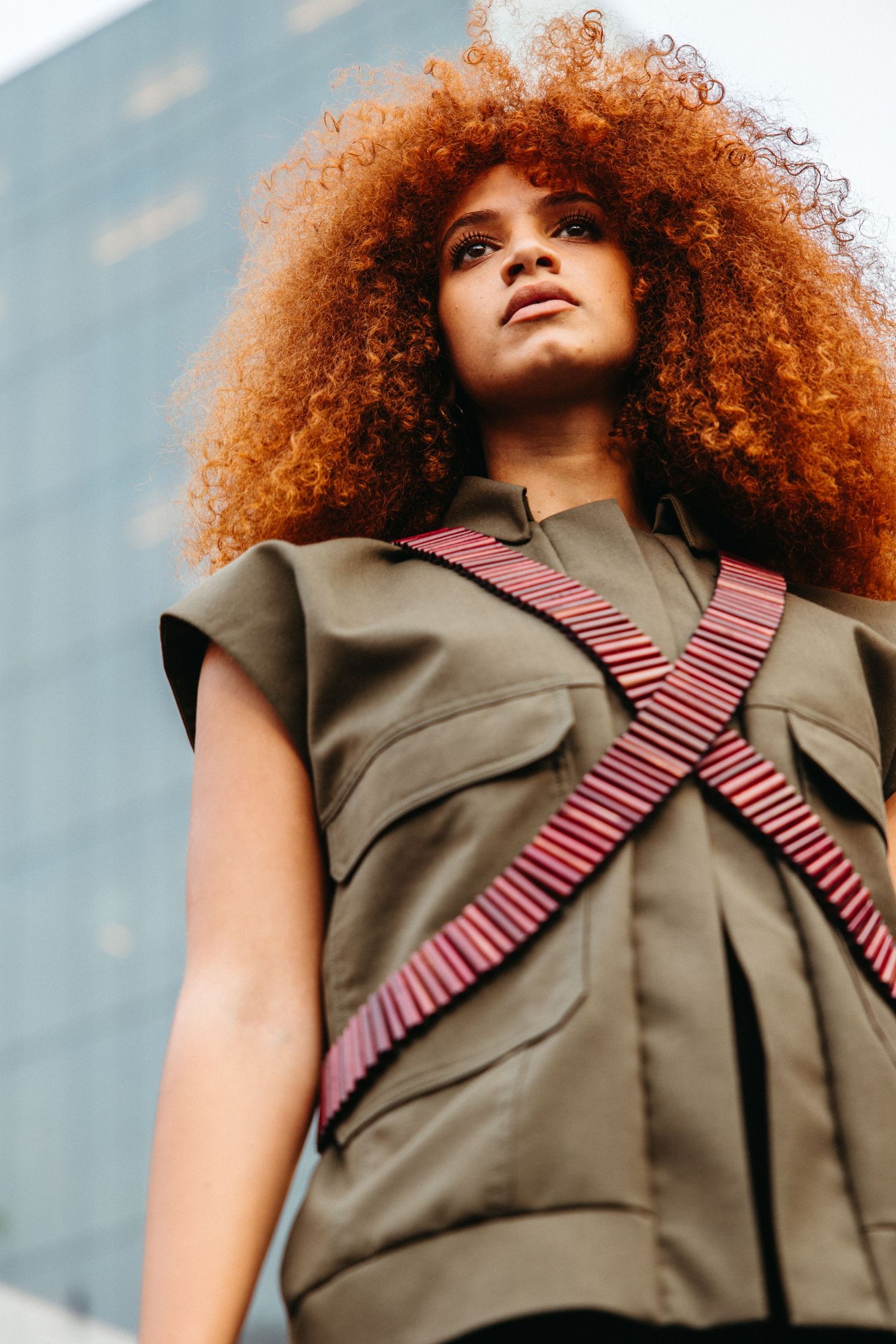 Low angle view of a person in olive green utility vest with red striped bandoliers and vibrant copper curly hair.