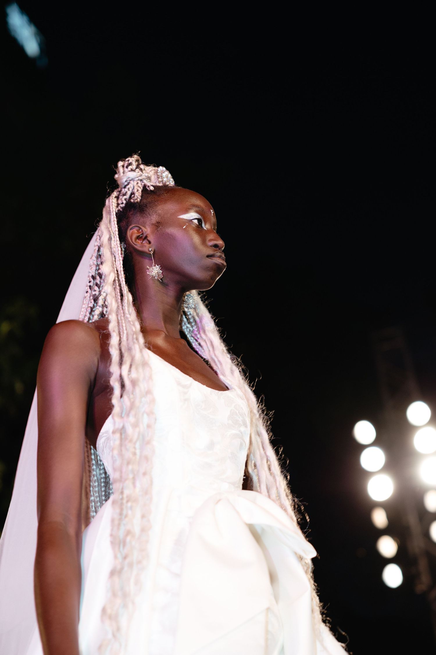 Model walks runway in elegant white bridal gown with long flowing veil and striking silver accessories.
