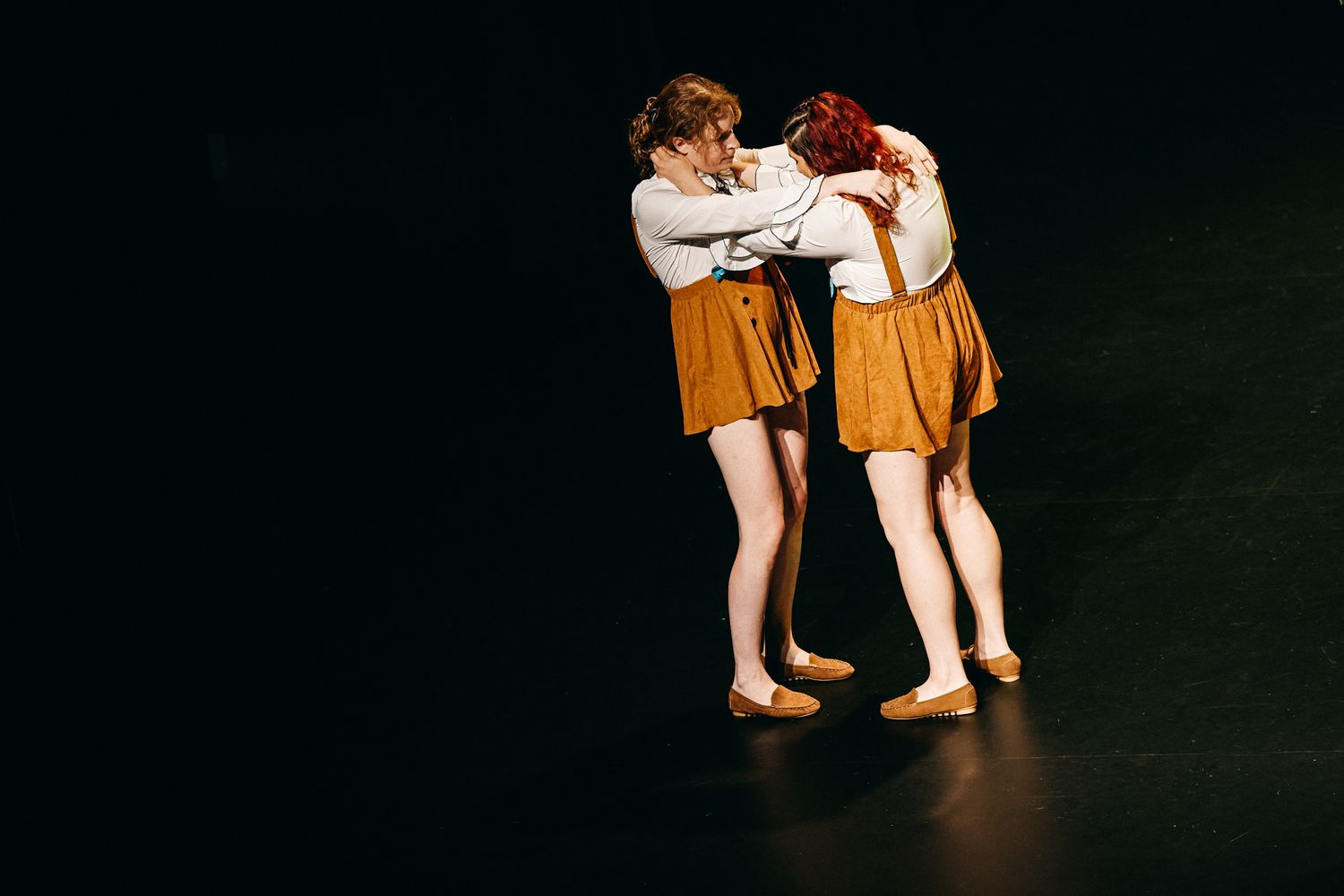 Two dancers in matching orange skirts embrace during a performance on a dark stage.