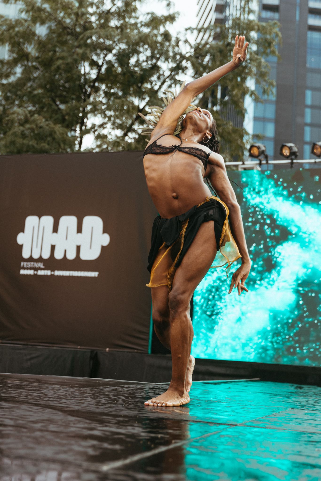 Dancer performs expressive contemporary dance movement on wet stage with turquoise lighting and urban backdrop.