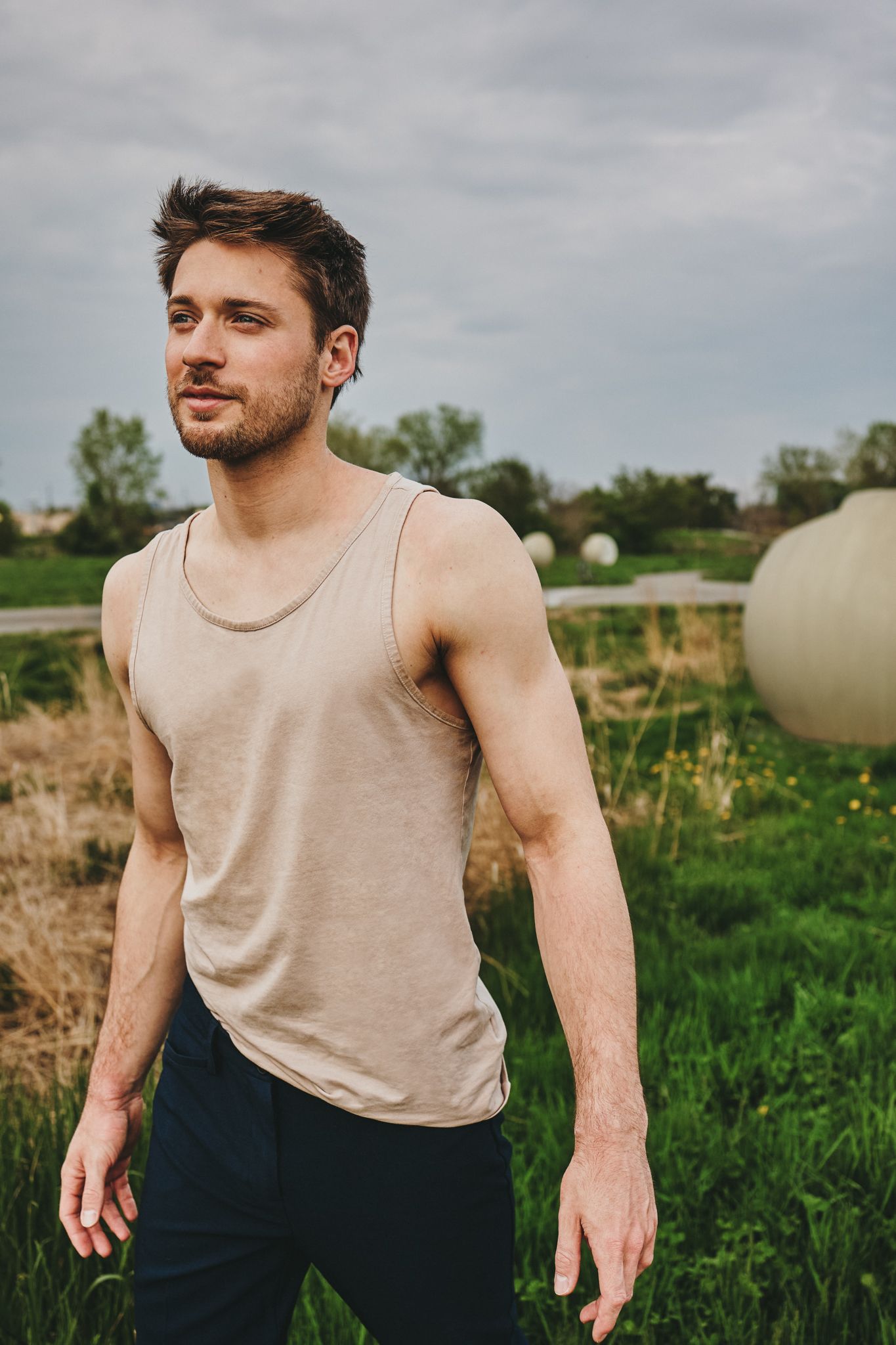 Athletic figure in beige tank top and dark pants moves dynamically through a green field with cloudy sky backdrop.