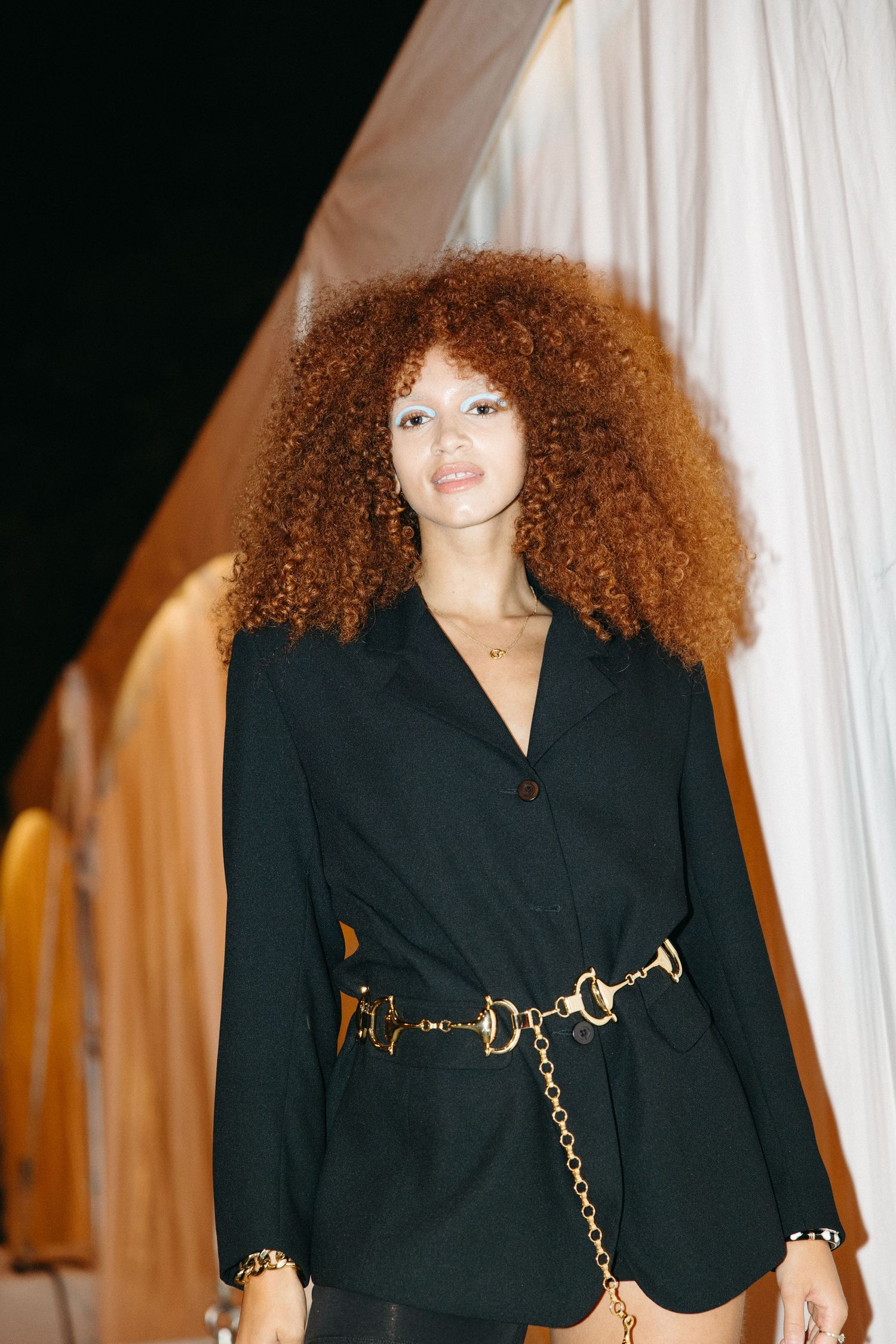 Model wearing a black blazer with gold chain belt poses against white backdrop in fashion photoshoot series.
