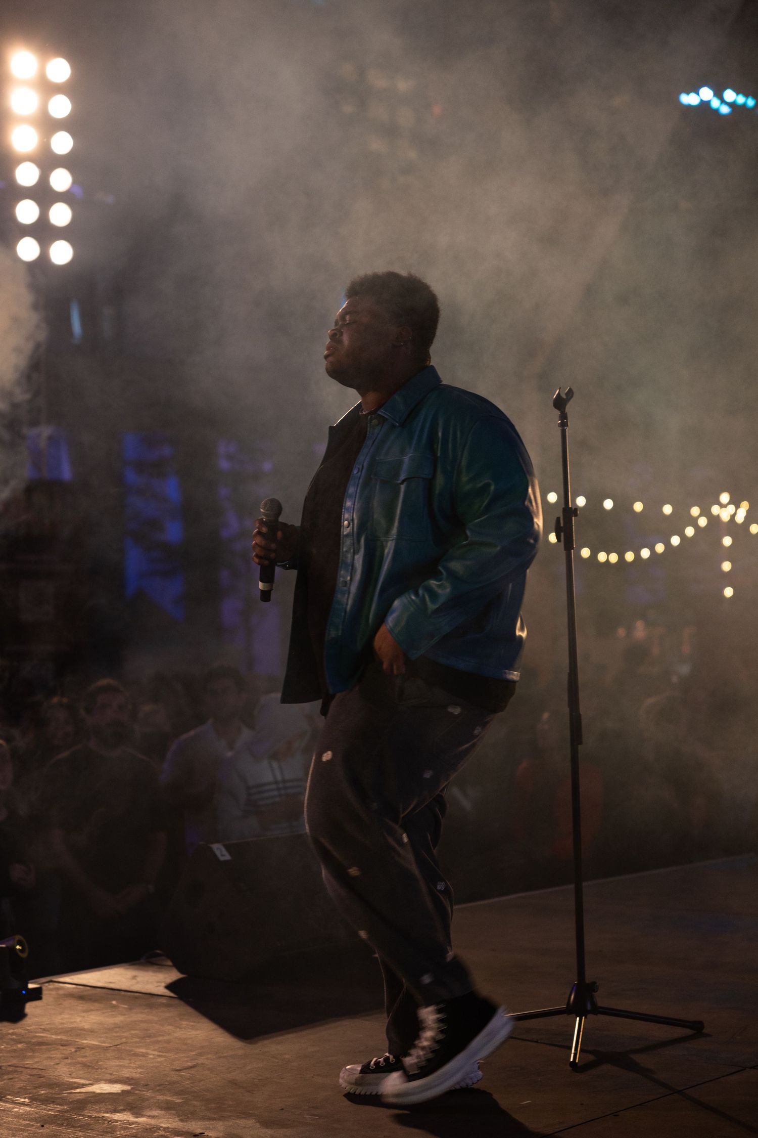 A performer in a blue jacket is silhouetted on a smoky concert stage with spotlights and string lights in the background.