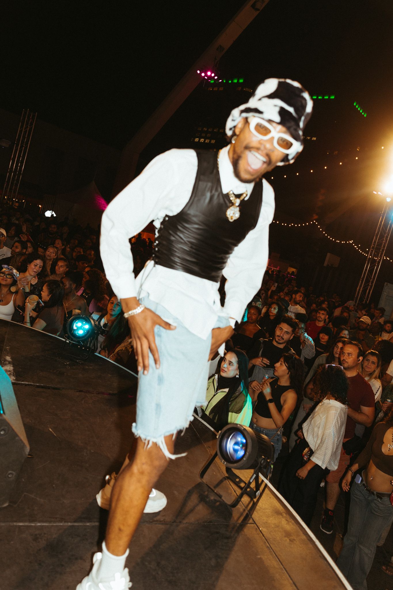 A performer on stage wears a white outfit with black vest and goggles while dancing energetically in front of a crowd.