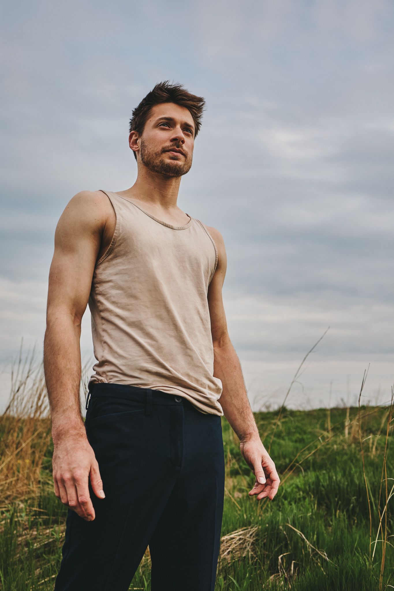 Person in beige tank top poses outdoors against cloudy sky and grassy field.