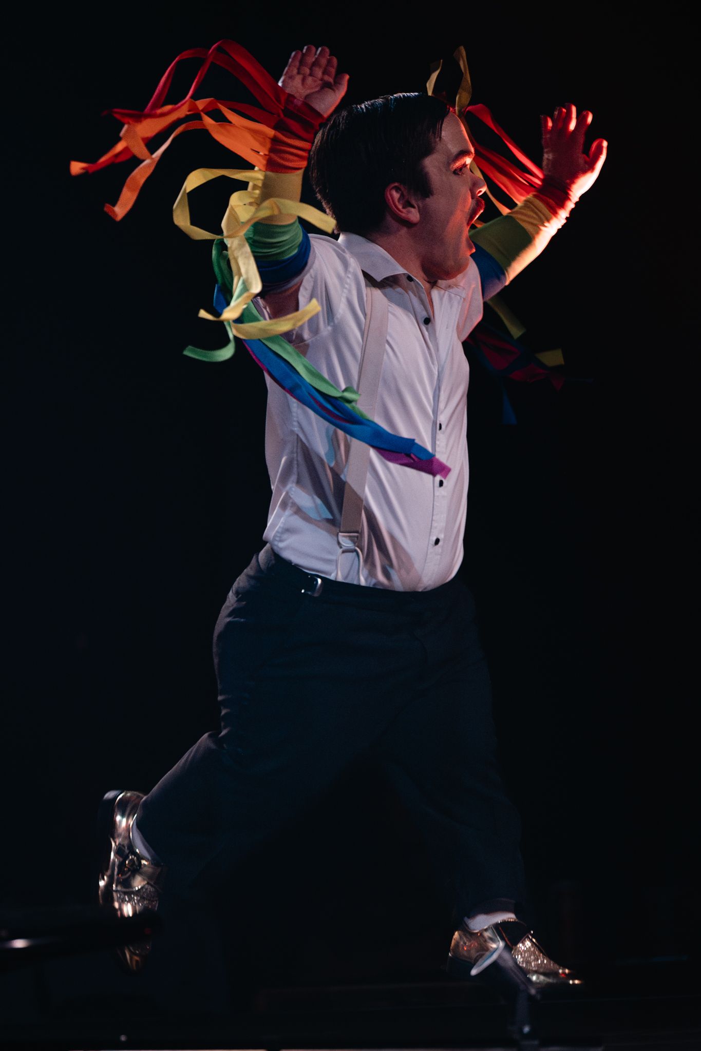 Dynamic stage performance with colorful rainbow ribbons flowing behind dancer in dress shirt and dark pants.
